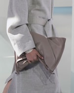 Placeholder for: Person in a light gray coat holds a large, soft taupe clutch handbag, with a blurred light background.