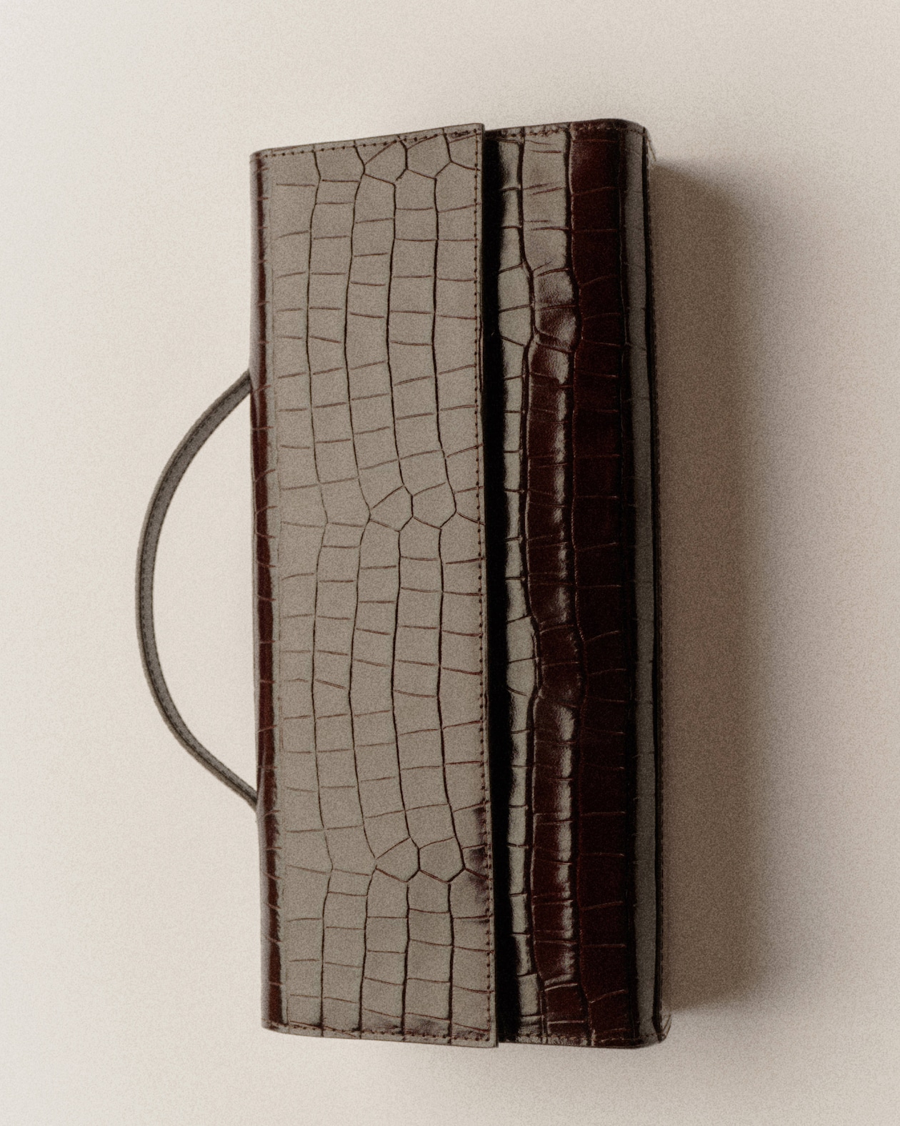 A rectangular, brown crocodile-patterned leather clutch with a handle, placed on a light surface.