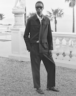 Placeholder for: Person in sunglasses wearing a dark suit and sandals, standing near a statue and balustrade with palm trees and the sea in the background.