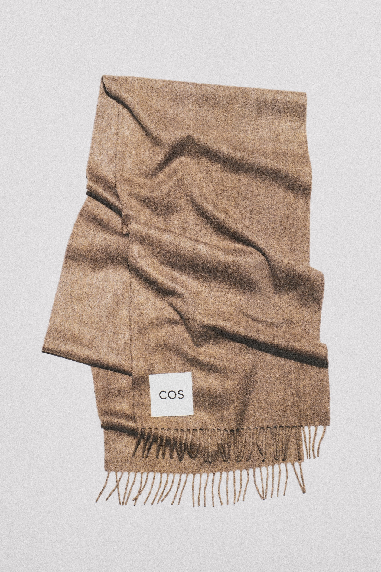 Folded tan wool scarf with fringe and white COS label on a light background.
