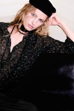 Placeholder for: Person in a sheer patterned blouse and black beret, resting their head on their hand, with a neutral expression.