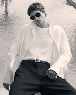 Placeholder for: Person in sunglasses and a white shirt leans back by a reflective water surface, exuding a relaxed, stylish demeanor. Black and white photo.