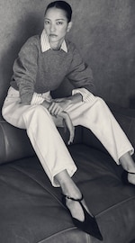 Placeholder for: A person with a serious expression sits on a couch, wearing a gray sweater over a striped shirt, white pants, and black shoes.
