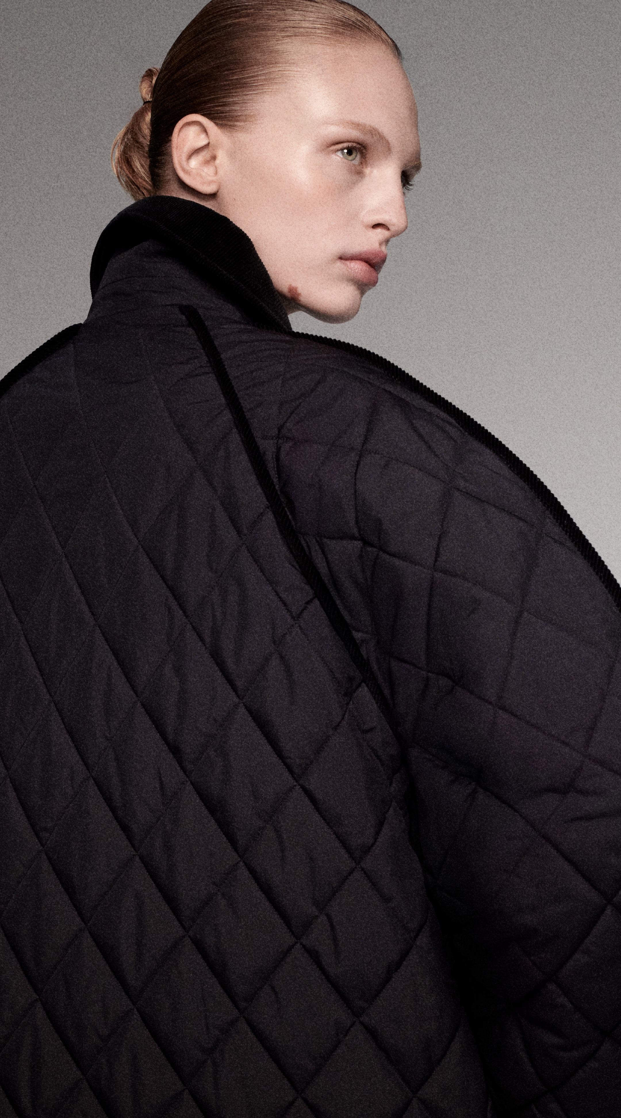 Person with slicked-back hair wearing a dark quilted jacket, looking off to the side against a neutral gray background.