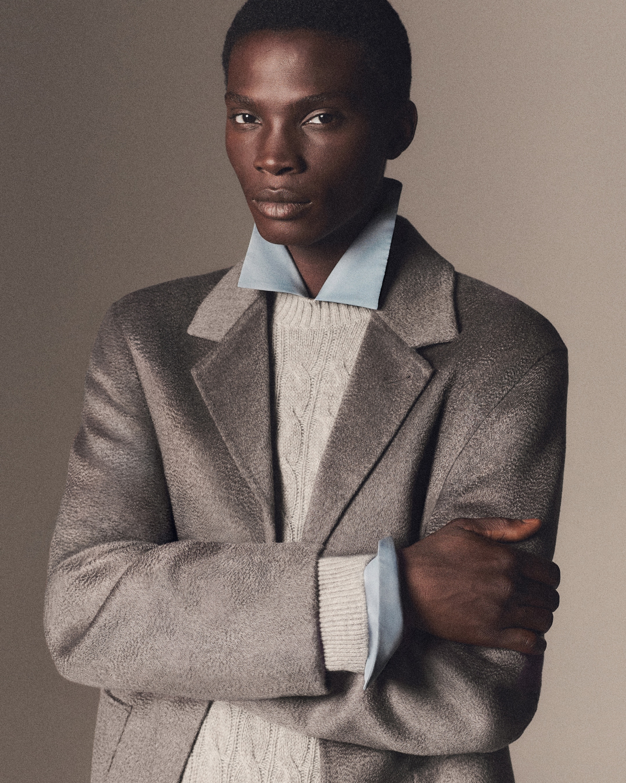 Person wearing a gray coat over a cable-knit sweater with a light blue shirt, posed with arms crossed against a neutral background.