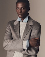 Placeholder for: Person wearing a gray coat over a cable-knit sweater with a light blue shirt, posed with arms crossed against a neutral background.