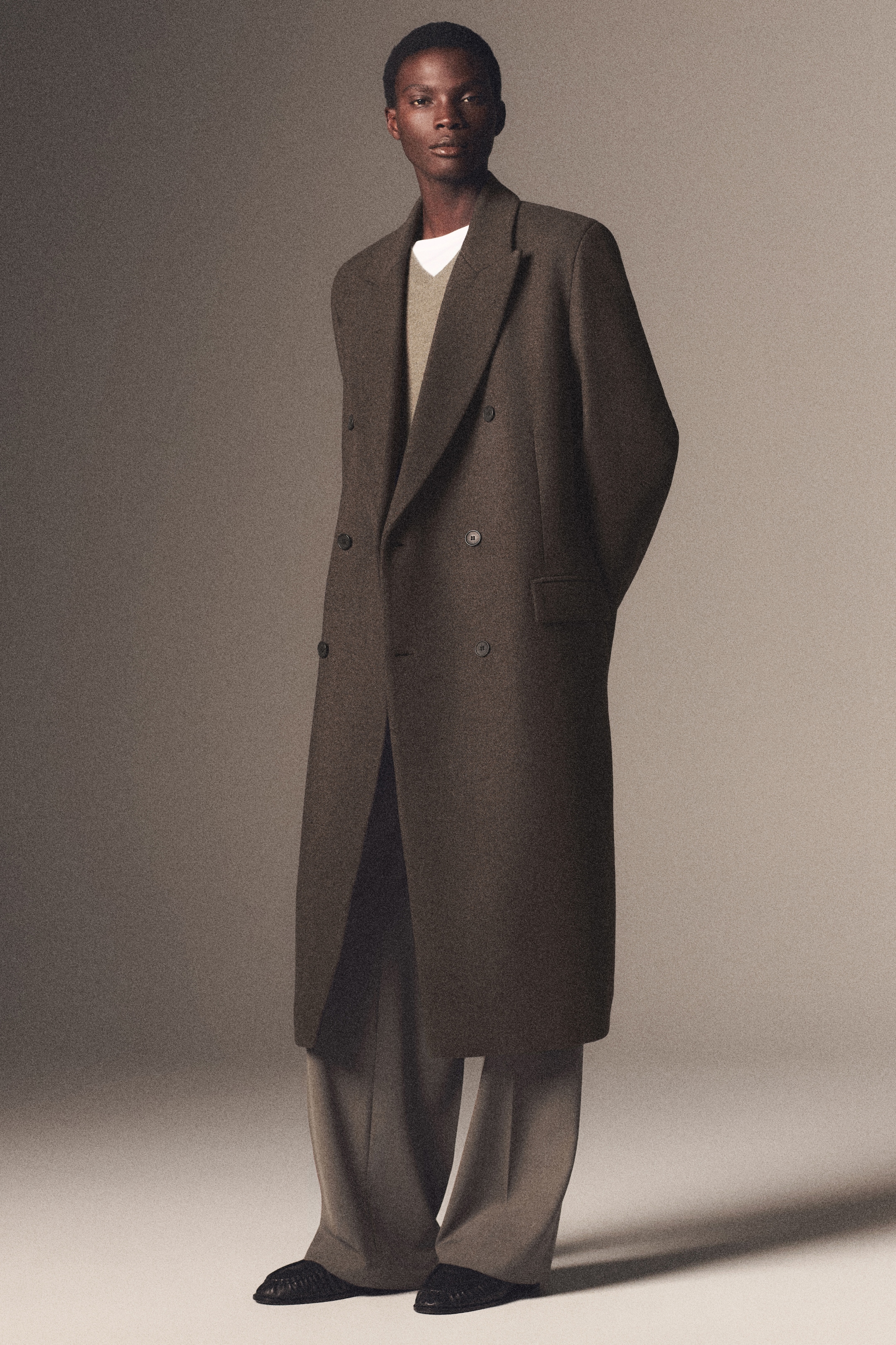 A person wearing a long, dark double-breasted coat with wide lapels, a light sweater, and trousers, standing against a neutral background.