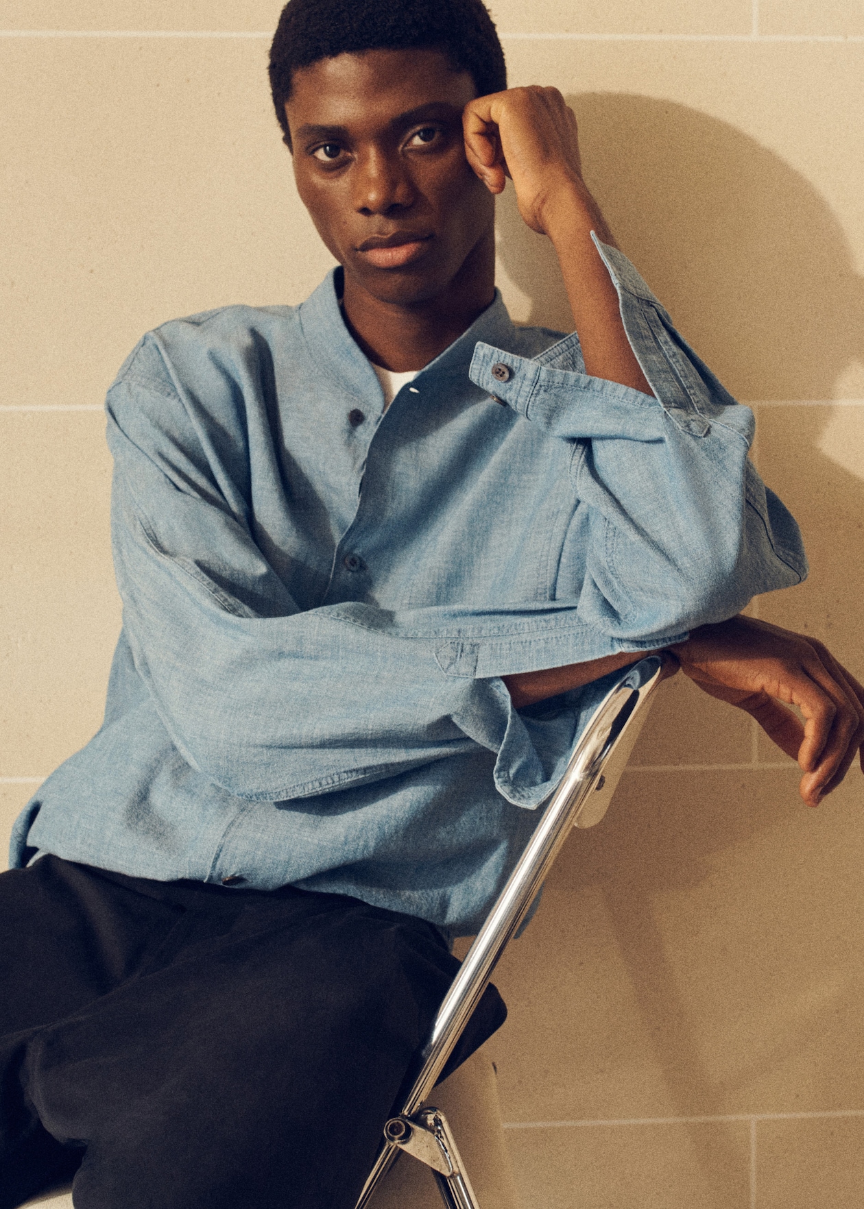 A person in a light blue button-up shirt sits on a metallic chair against a beige tiled wall, looking thoughtful.