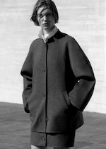 Placeholder for: Black and white photo of a person wearing a large, buttoned coat and standing against a textured wall with hands in pockets.