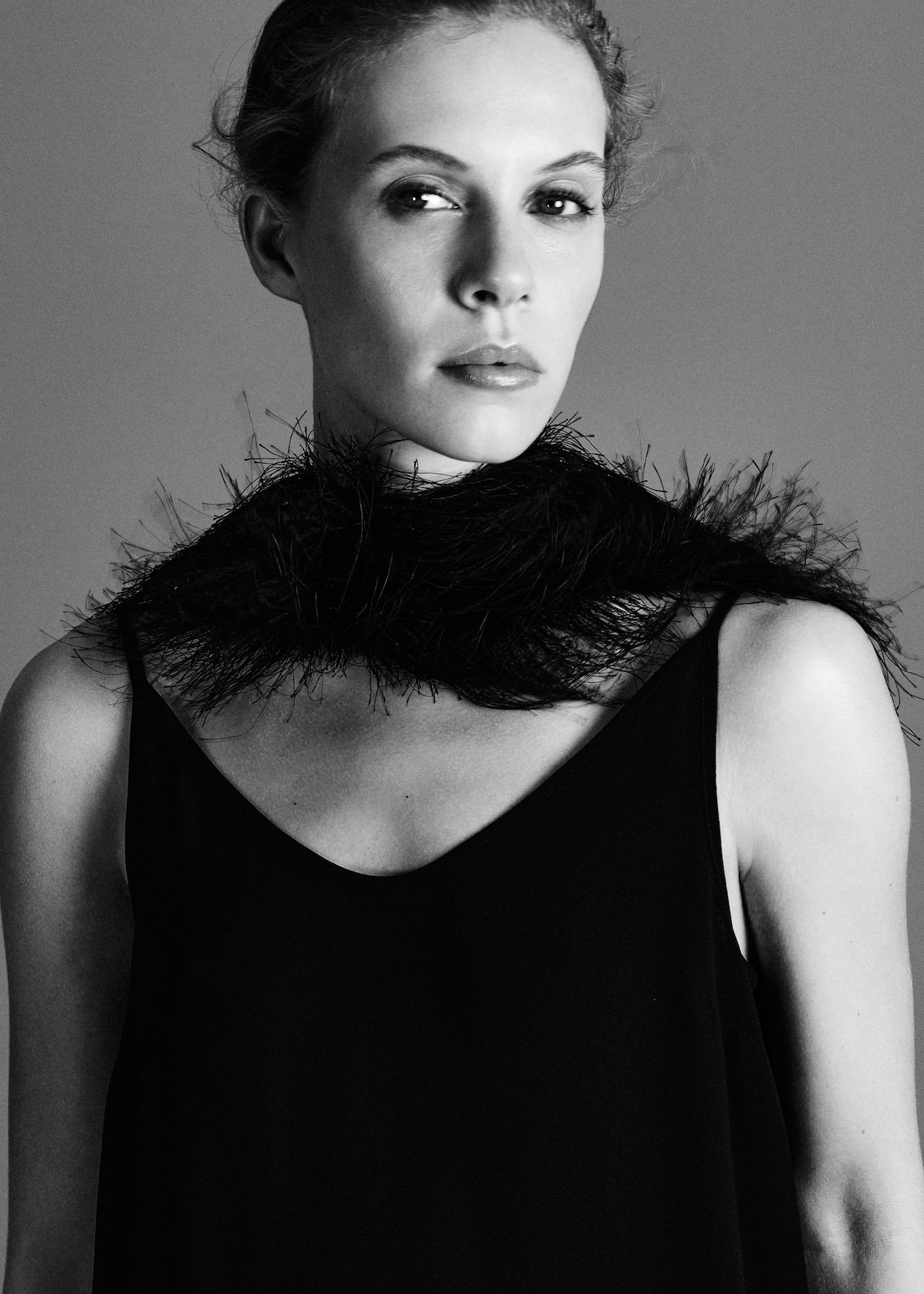 Black and white portrait of a woman wearing a dark sleeveless dress and a feathered scarf, looking at the camera with a neutral expression.