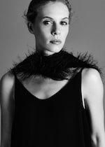 Placeholder for: Black and white portrait of a woman wearing a dark sleeveless dress and a feathered scarf, looking at the camera with a neutral expression.