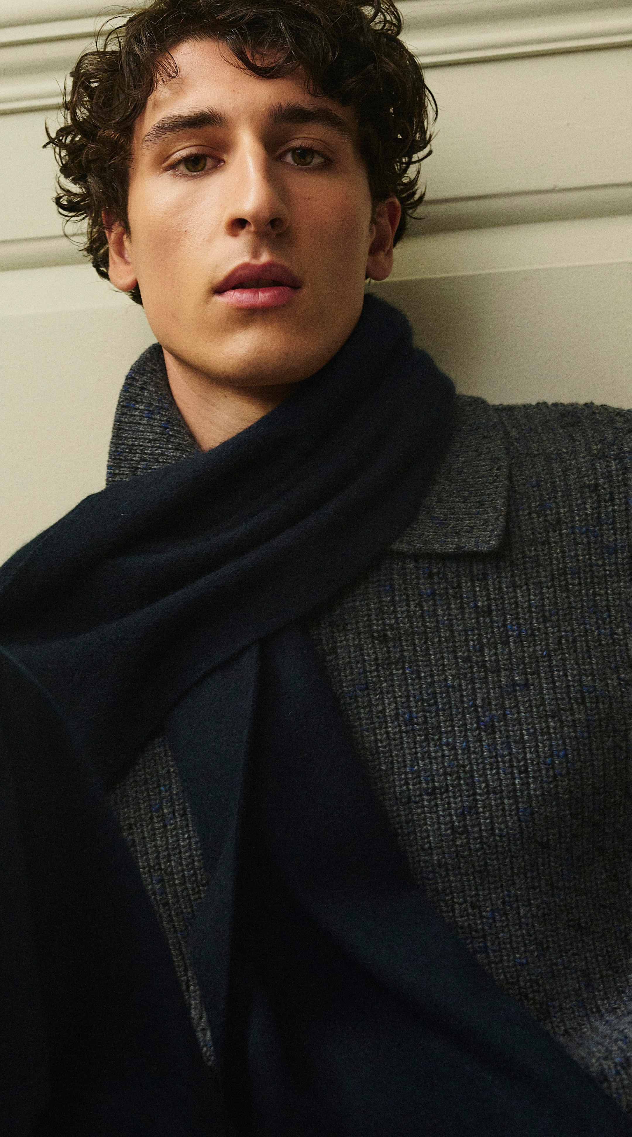 Person with curly hair wearing a navy scarf and gray knit sweater, looking directly at the camera against a light-colored wall.