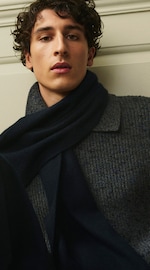 Placeholder for: Person with curly hair wearing a navy scarf and gray knit sweater, looking directly at the camera against a light-colored wall.