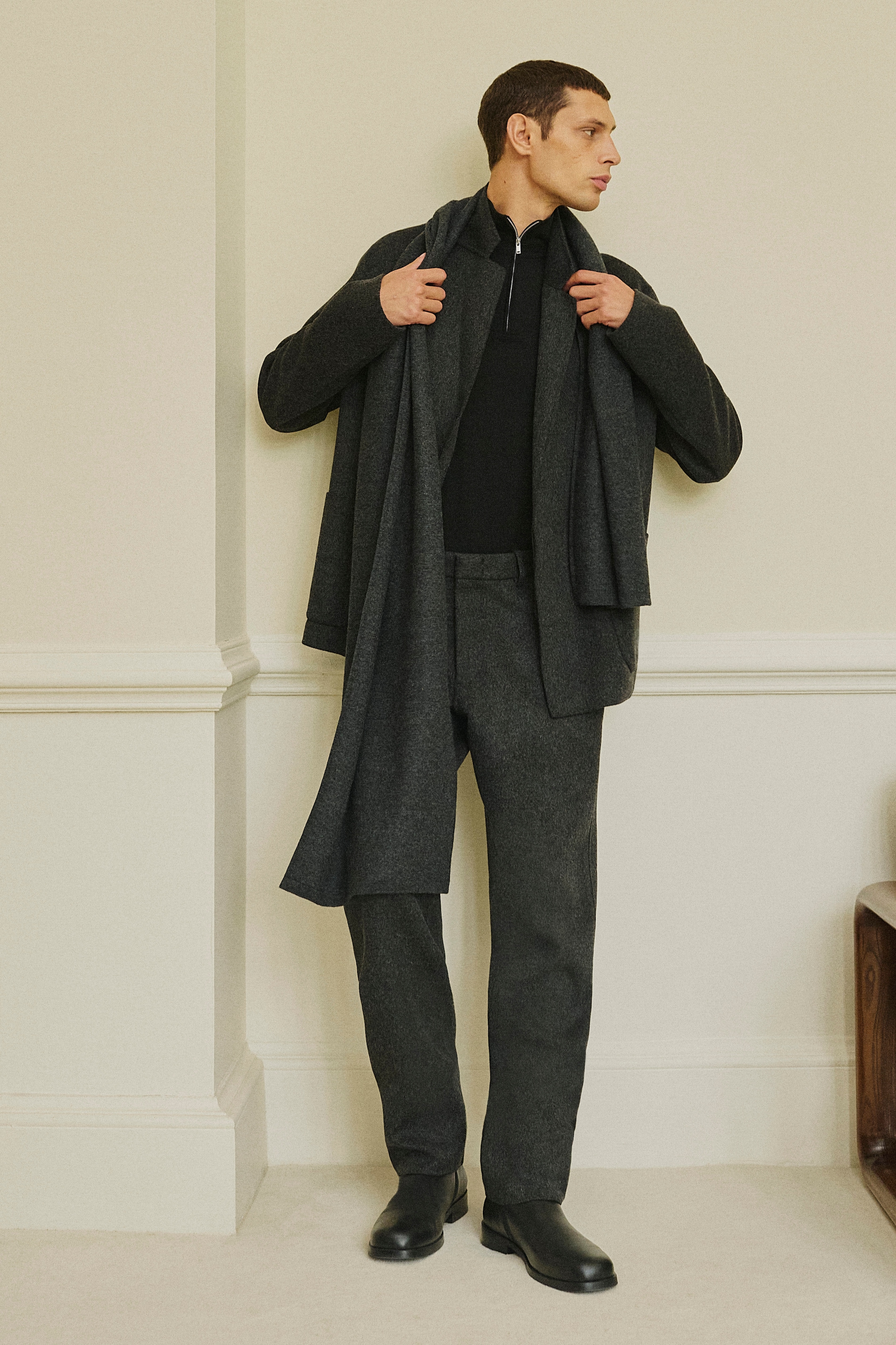 A man in a dark outfit stands against a light wall. He's wearing a coat, scarf, and trousers, with black shoes, looking to the side.