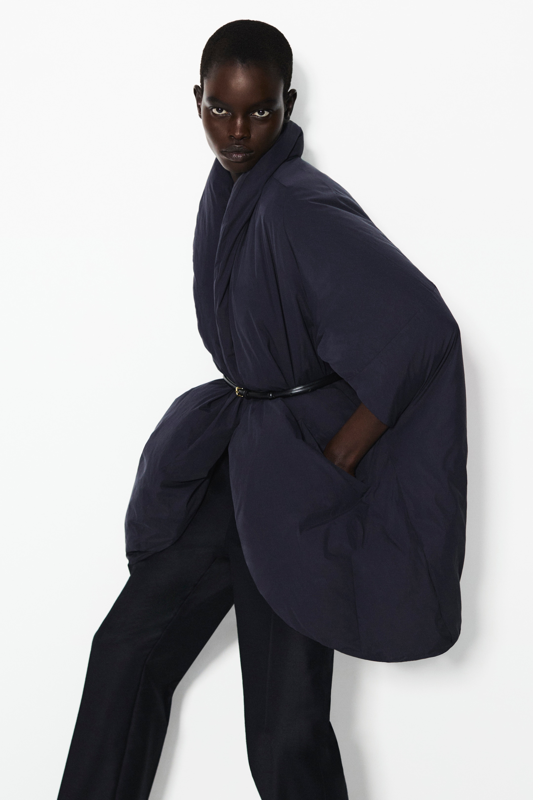 Person in a dark blue, oversized kimono-style coat, belted at the waist, with hands in pockets, standing against a white background.