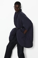 Placeholder for: Person in a dark blue, oversized kimono-style coat, belted at the waist, with hands in pockets, standing against a white background.
