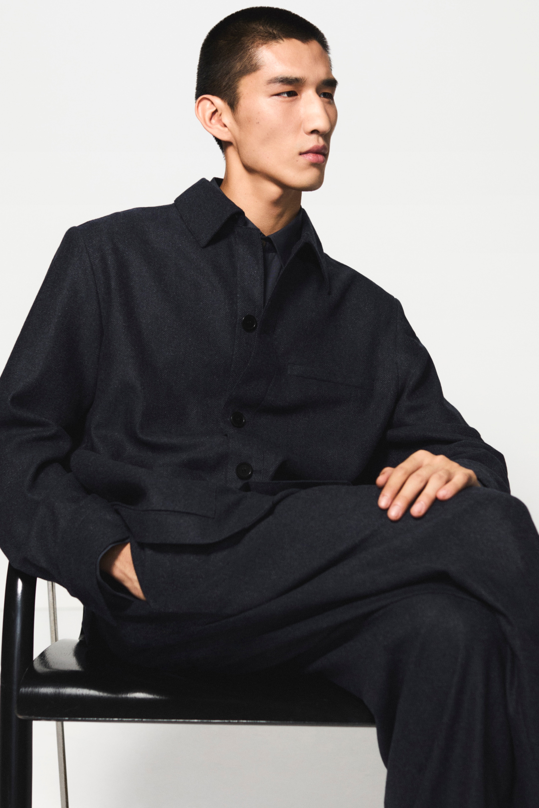 Person in a dark suit sits on a chair with legs crossed, looking to the side against a plain white background.