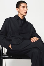 Placeholder for: Person in a dark suit sits on a chair with legs crossed, looking to the side against a plain white background.