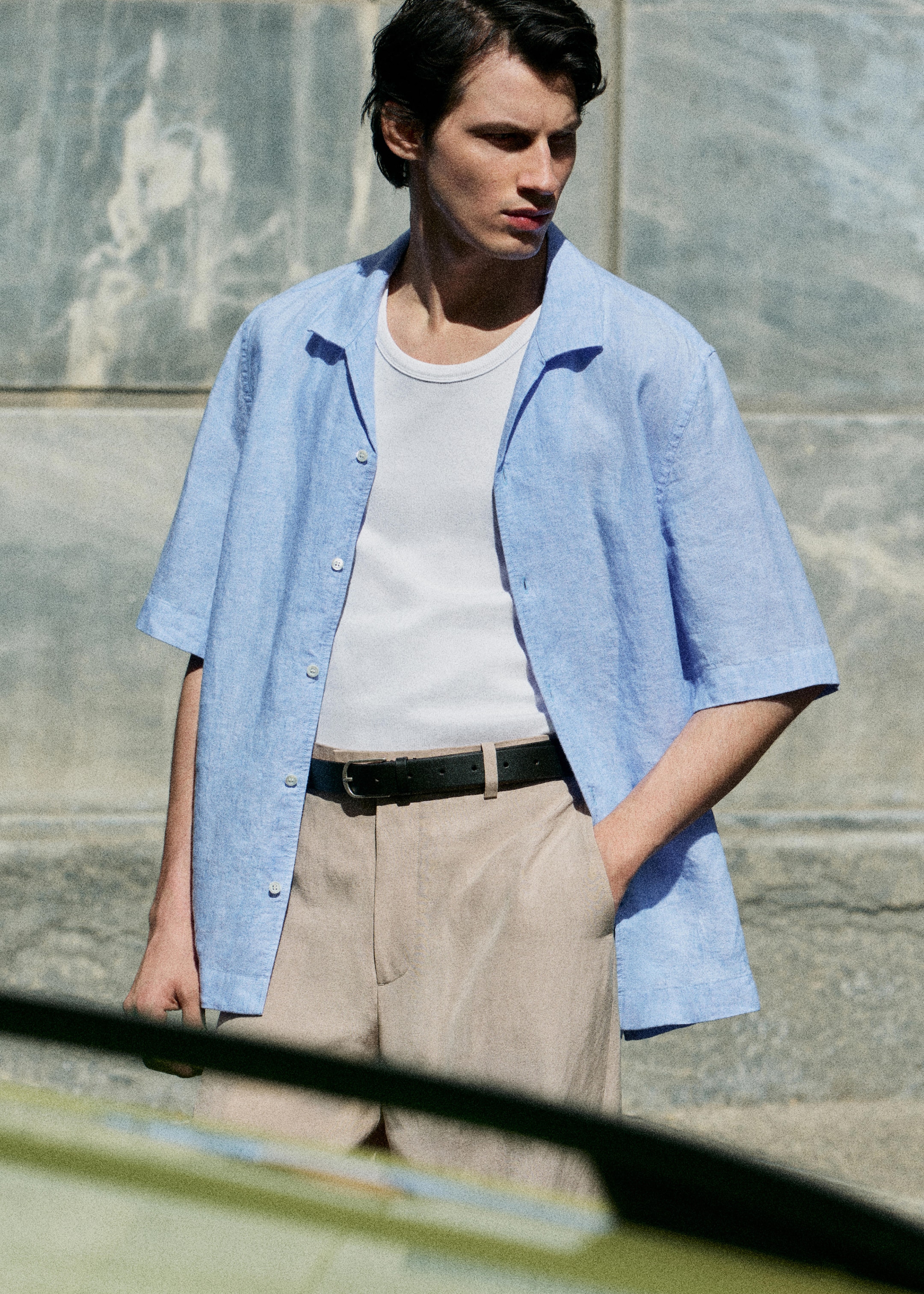 Man in a light blue shirt, white tank top, and beige pants, standing outdoors with one hand in pocket, looking to the side.
