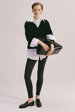 Placeholder for: Person in a black sweater over a white shirt, black leggings, and loafers, holding a large black bag against a plain background.