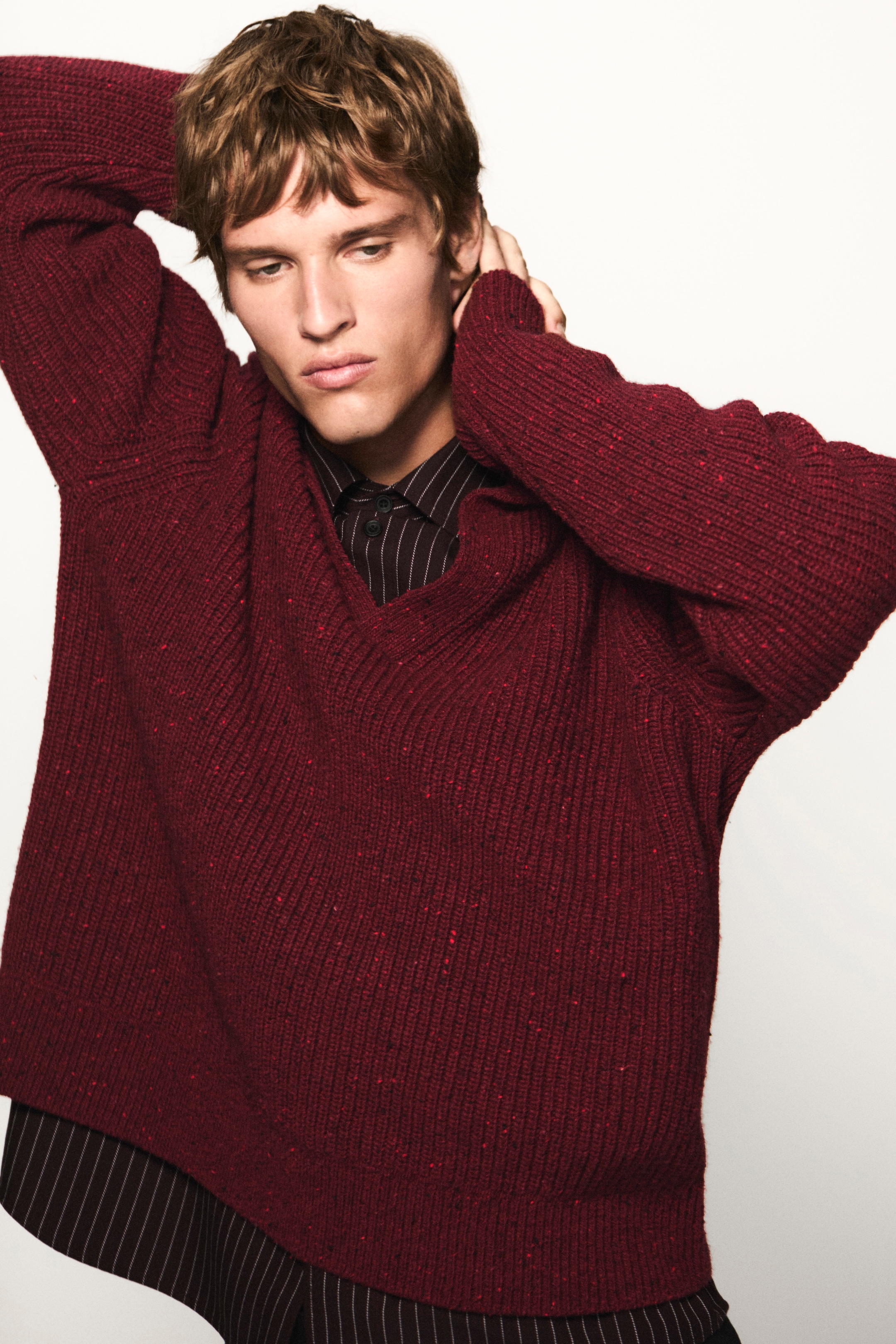 Person in a maroon sweater over a pinstriped shirt, posing with arms raised against a plain background.