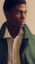 Placeholder for: A person wearing a green jacket over a striped button-up shirt, looking thoughtfully to the side against a neutral background.