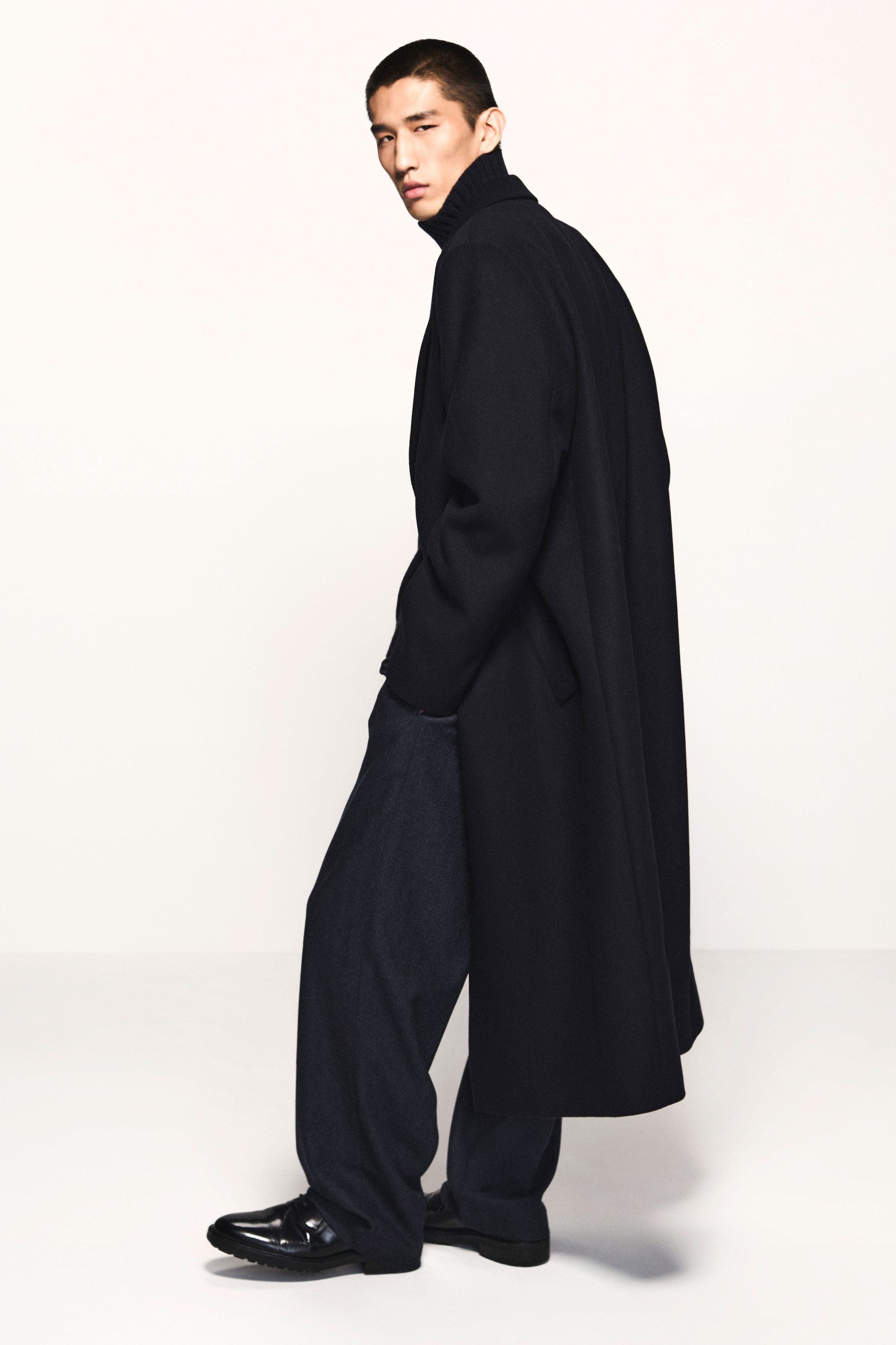 Person wearing a long, dark coat and wide-legged pants, standing against a plain background, looking to the side.