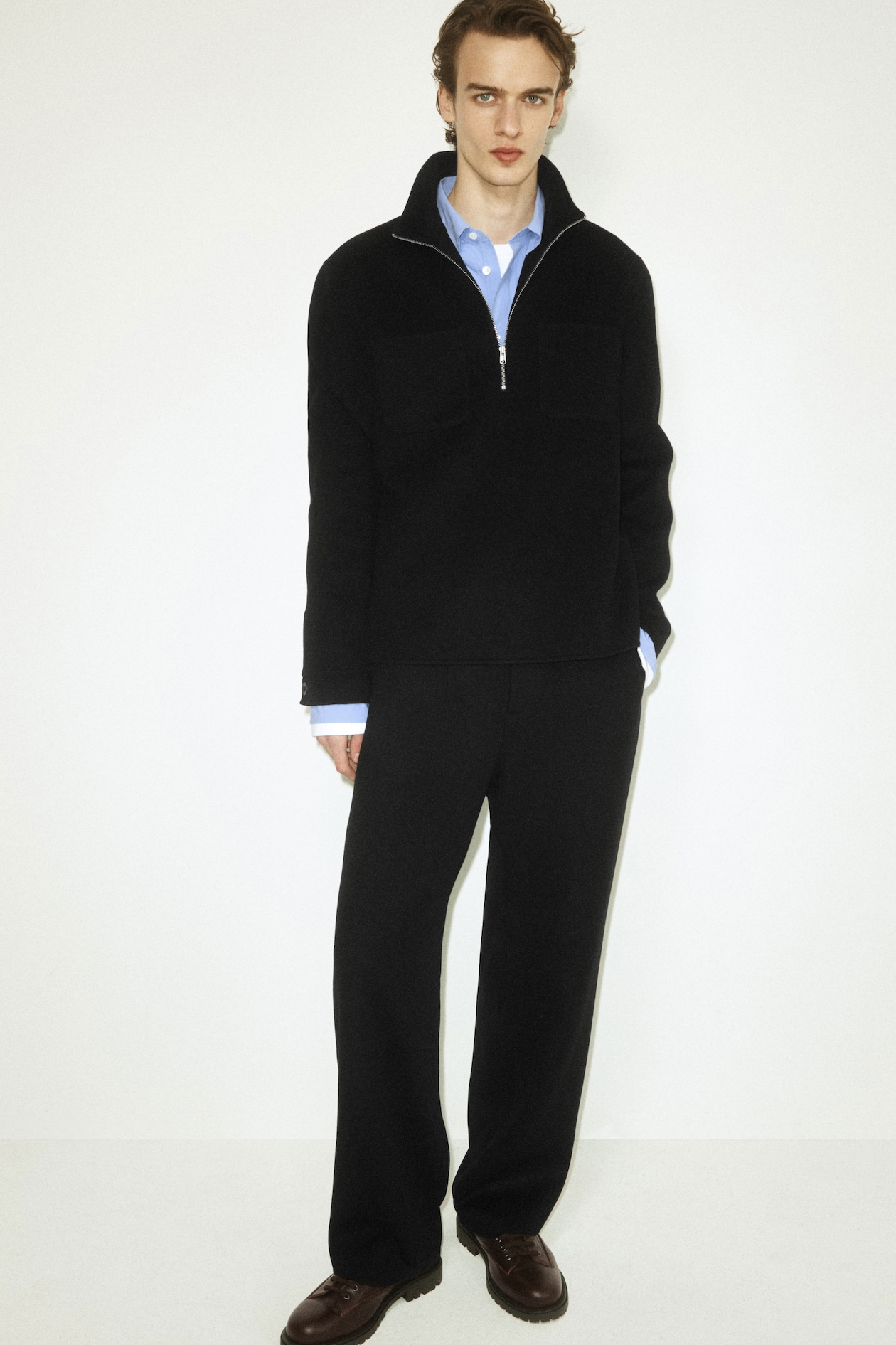 Person wearing a black zip-up sweater, blue shirt, black pants, and brown shoes, standing against a plain white background.