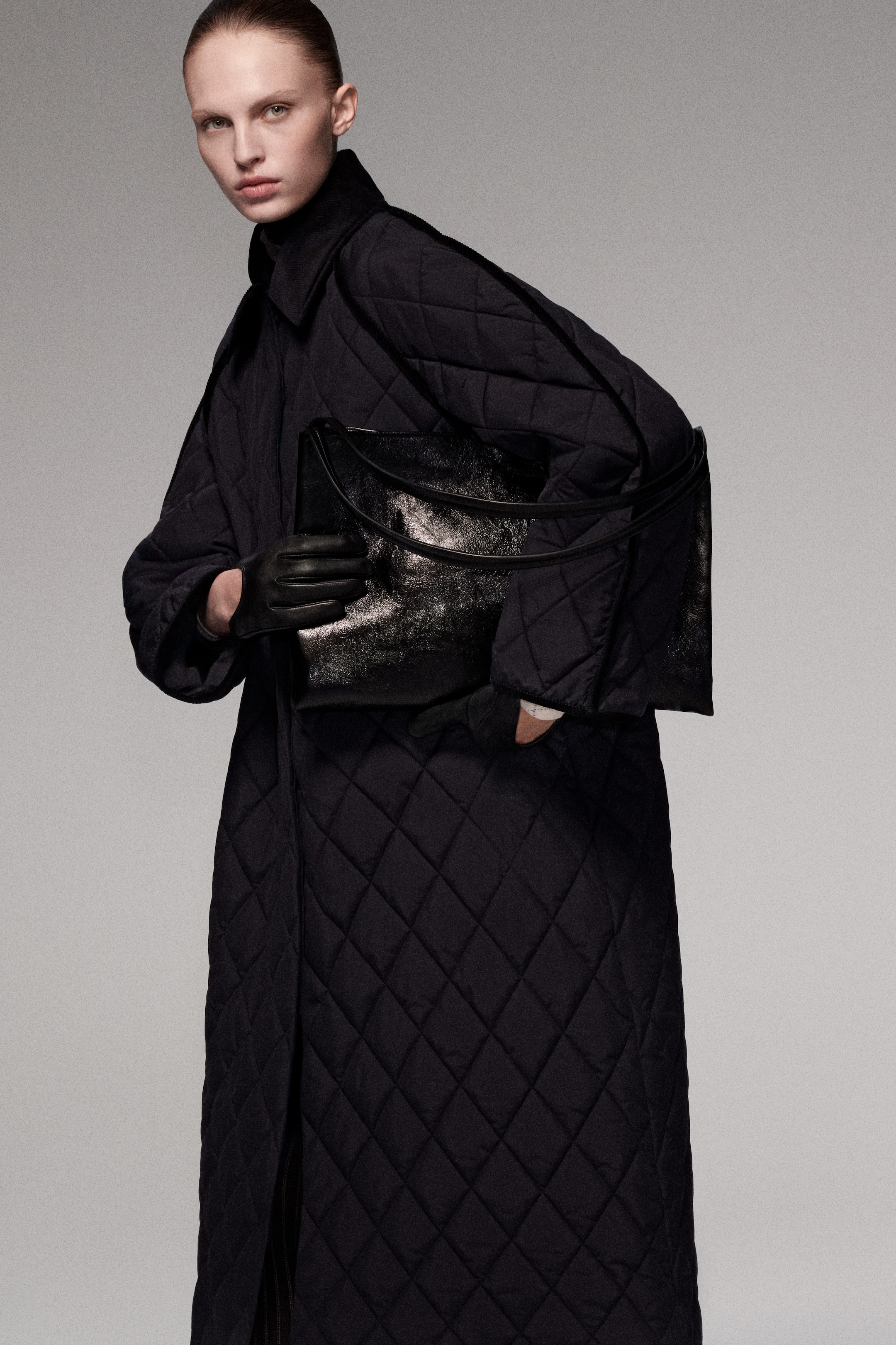 Person wearing a long quilted coat, holding a large black leather bag, with dark gloves, against a plain background.