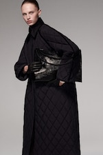 Placeholder for: Person wearing a long quilted coat, holding a large black leather bag, with dark gloves, against a plain background.