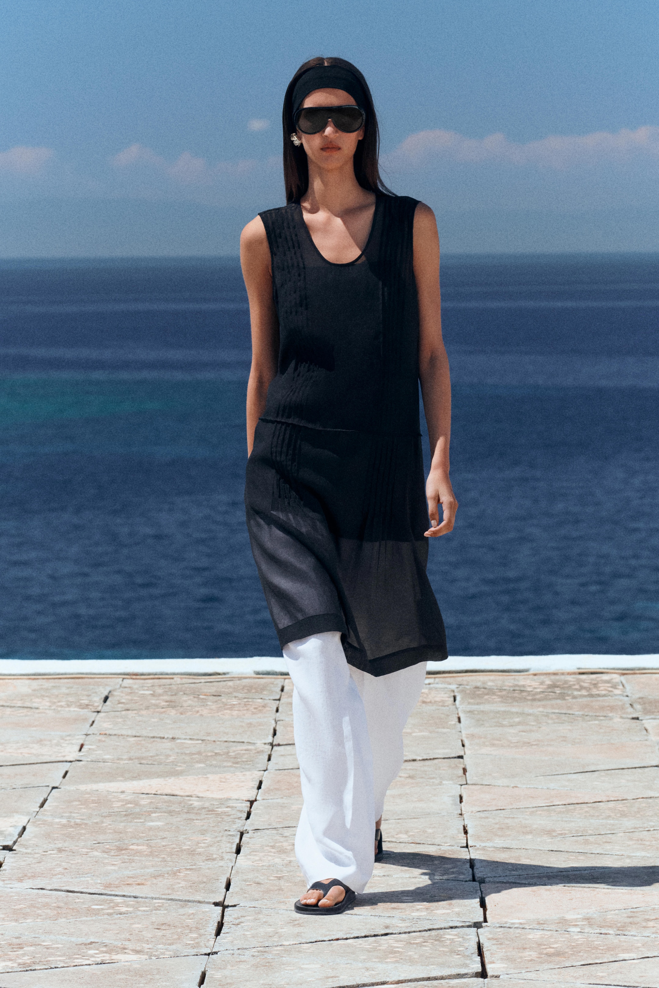 A person in a sleeveless black tunic, white pants, sunglasses, and sandals walks on a stone path by a blue sea under a clear sky.