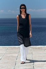Placeholder for: A person in a sleeveless black tunic, white pants, sunglasses, and sandals walks on a stone path by a blue sea under a clear sky.