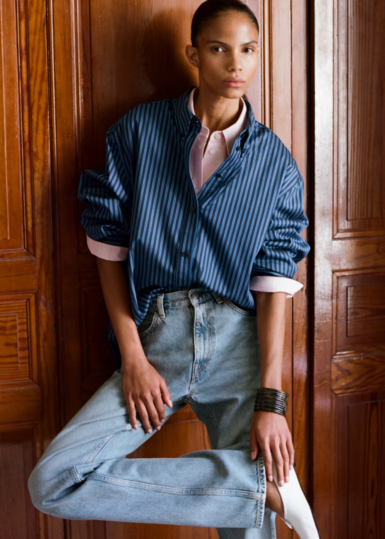 Person wearing a blue striped shirt, pink undershirt, light blue jeans, and white socks, leaning against a wooden wall.