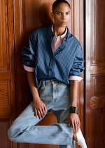 Placeholder for: Person wearing a blue striped shirt, pink undershirt, light blue jeans, and white socks, leaning against a wooden wall.