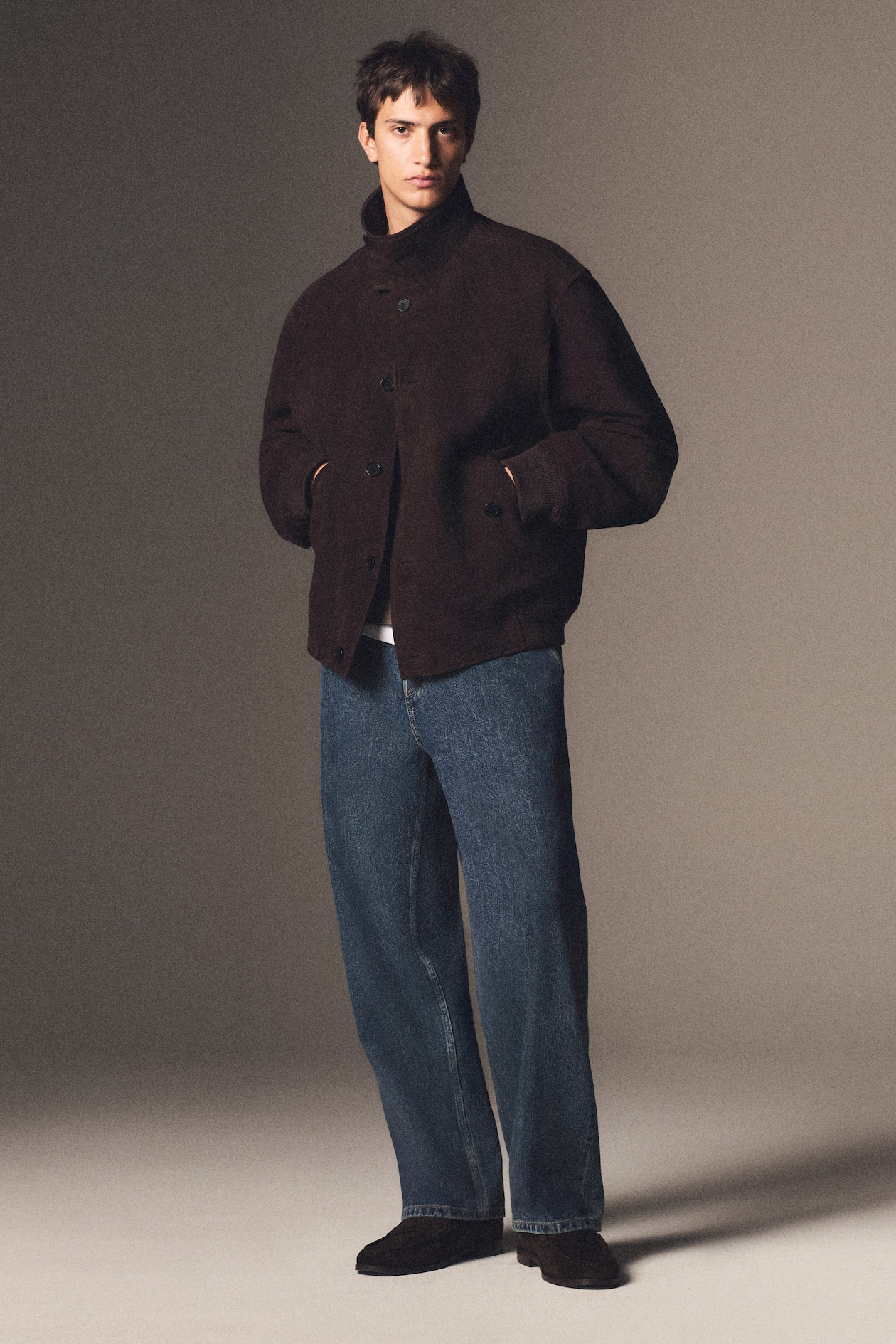 A man wearing a dark brown jacket, blue jeans, and black shoes stands against a neutral background.