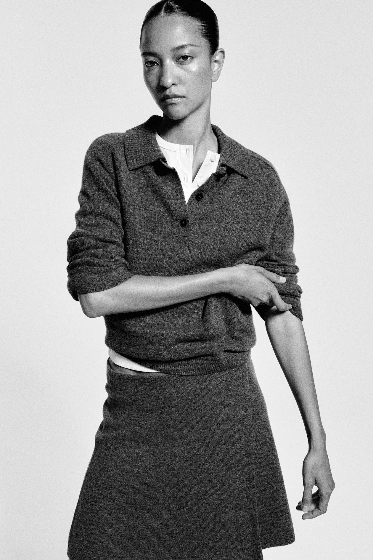 A person in black and white wearing a dark sweater and skirt, looking at the camera with a neutral expression, adjusting their sleeve.