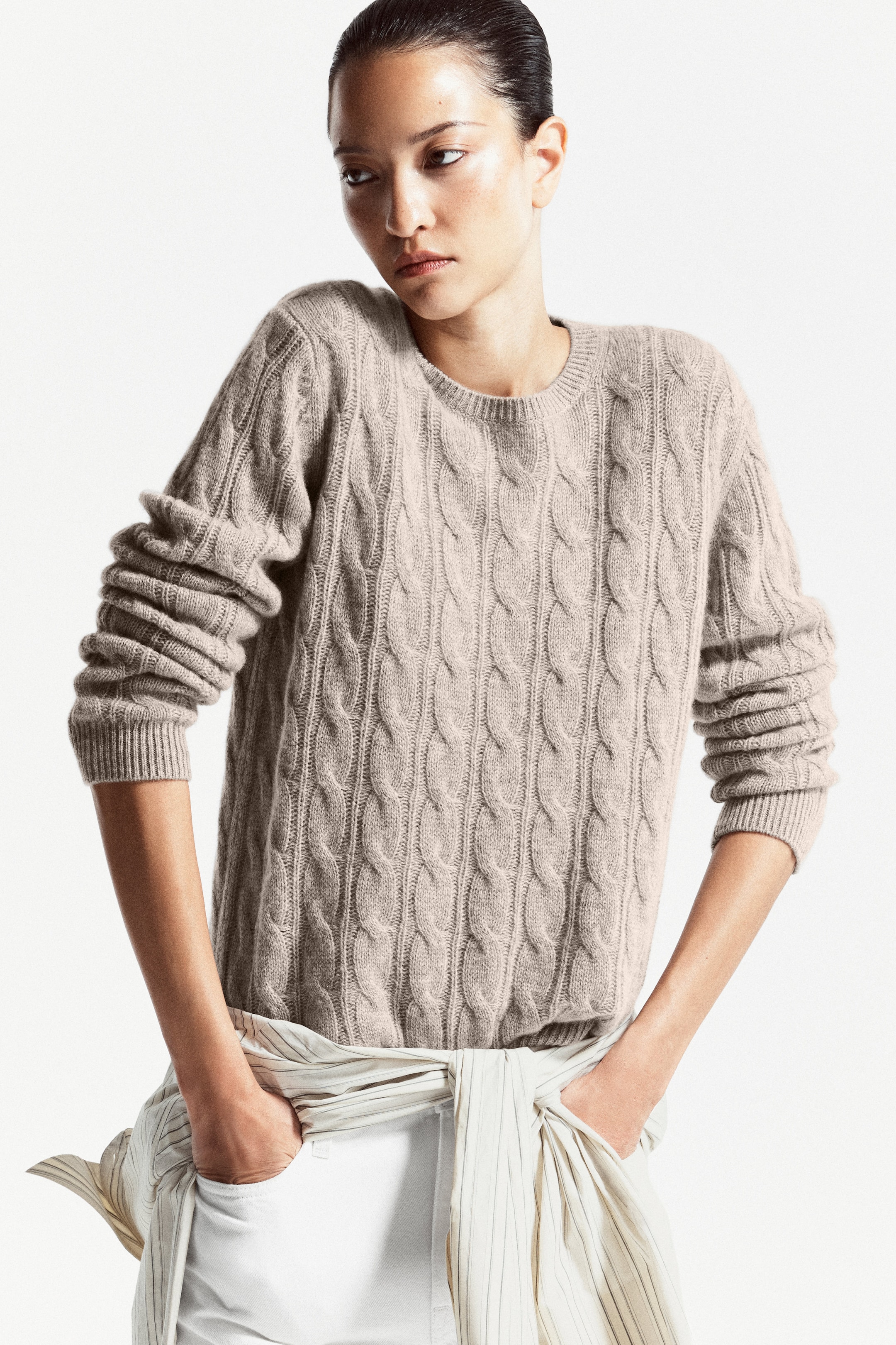 Person wearing a beige cable-knit sweater with rolled-up sleeves, standing against a white background, and looking to the side.