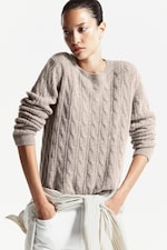 Placeholder for: Person wearing a beige cable-knit sweater with rolled-up sleeves, standing against a white background, and looking to the side.