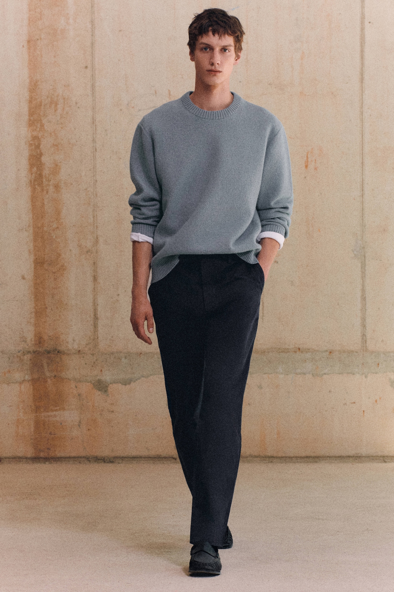 A person wearing a light gray sweater, white undershirt, and dark pants walks confidently against a textured beige wall background.