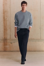 Placeholder for: A person wearing a light gray sweater, white undershirt, and dark pants walks confidently against a textured beige wall background.