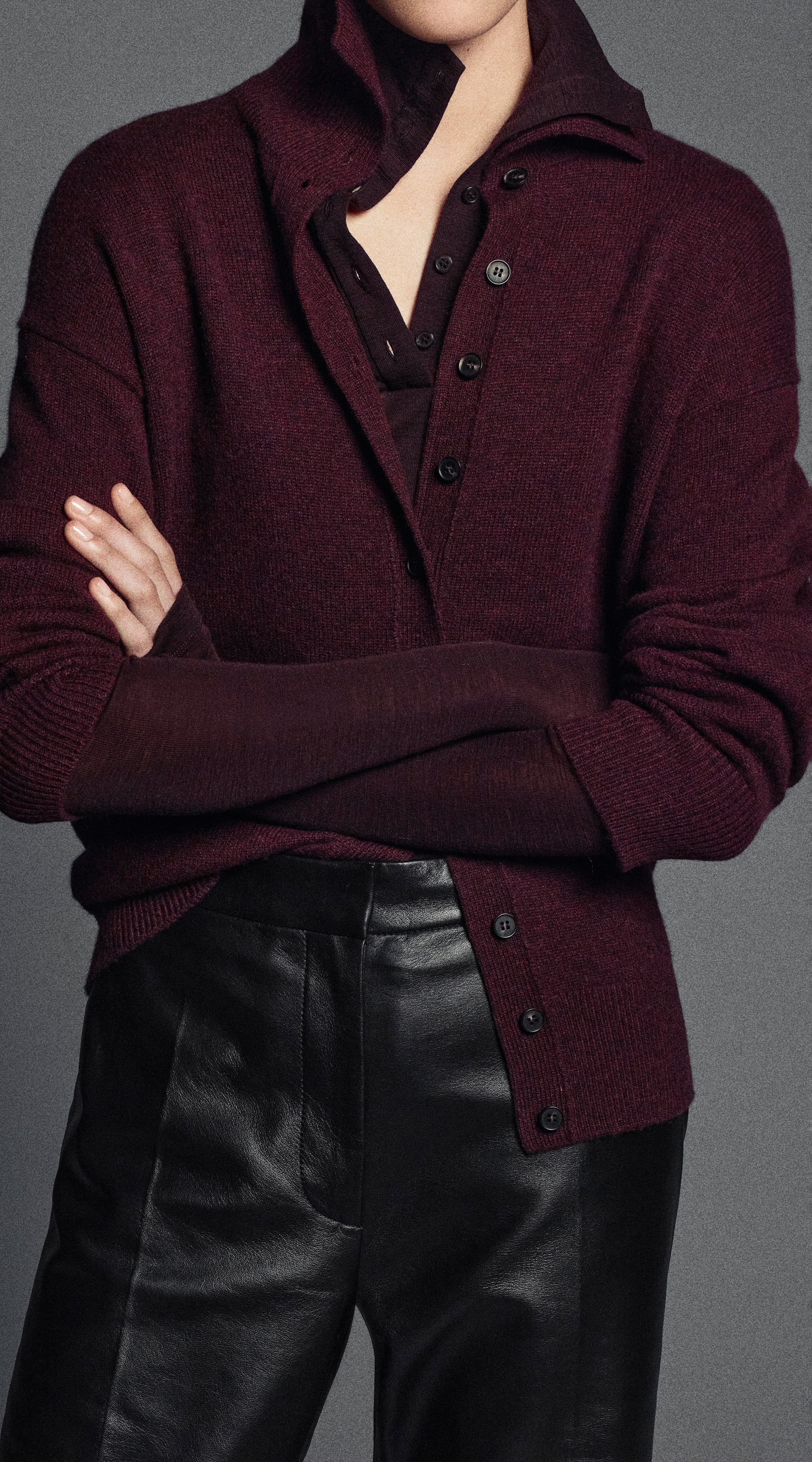 Woman in a burgundy button-up sweater and leather pants with arms crossed, shown from neck to waist against a neutral background.
