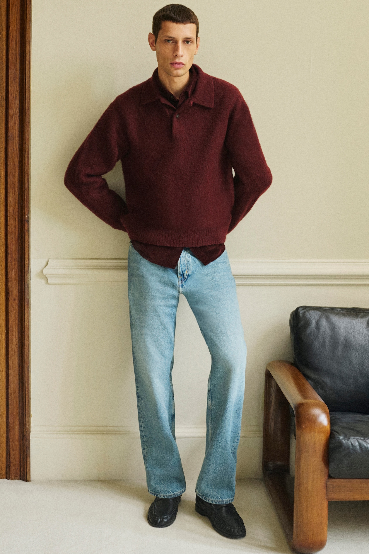 Man wearing a burgundy sweater and light blue jeans stands indoors with arms crossed, beside a black leather chair.