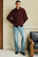 Placeholder for: Man wearing a burgundy sweater and light blue jeans stands indoors with arms crossed, beside a black leather chair.