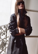 Placeholder for: Person in black leather coat and sunglasses, with long hair, standing with arms crossed near a decorative railing in a sunlit interior.