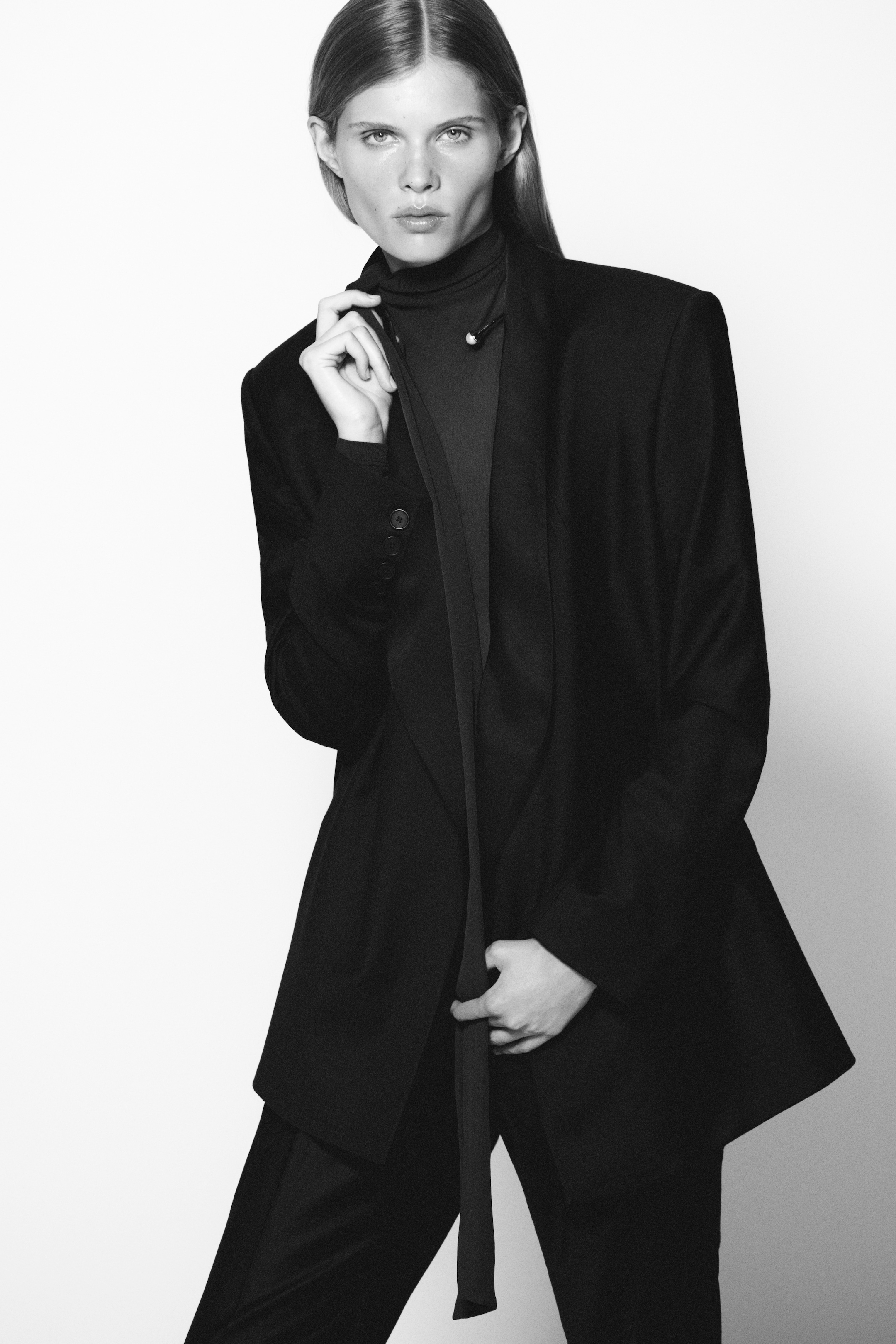 Person in a black suit and turtleneck, posing confidently against a plain background. The image is in black and white.