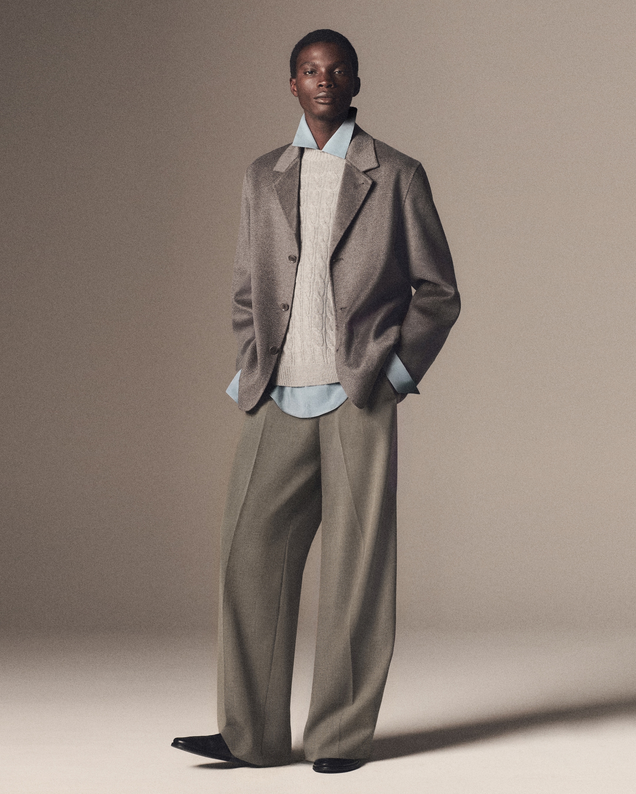 A person in a gray blazer, cable-knit sweater, and light blue shirt, with wide-legged trousers, standing against a neutral background.