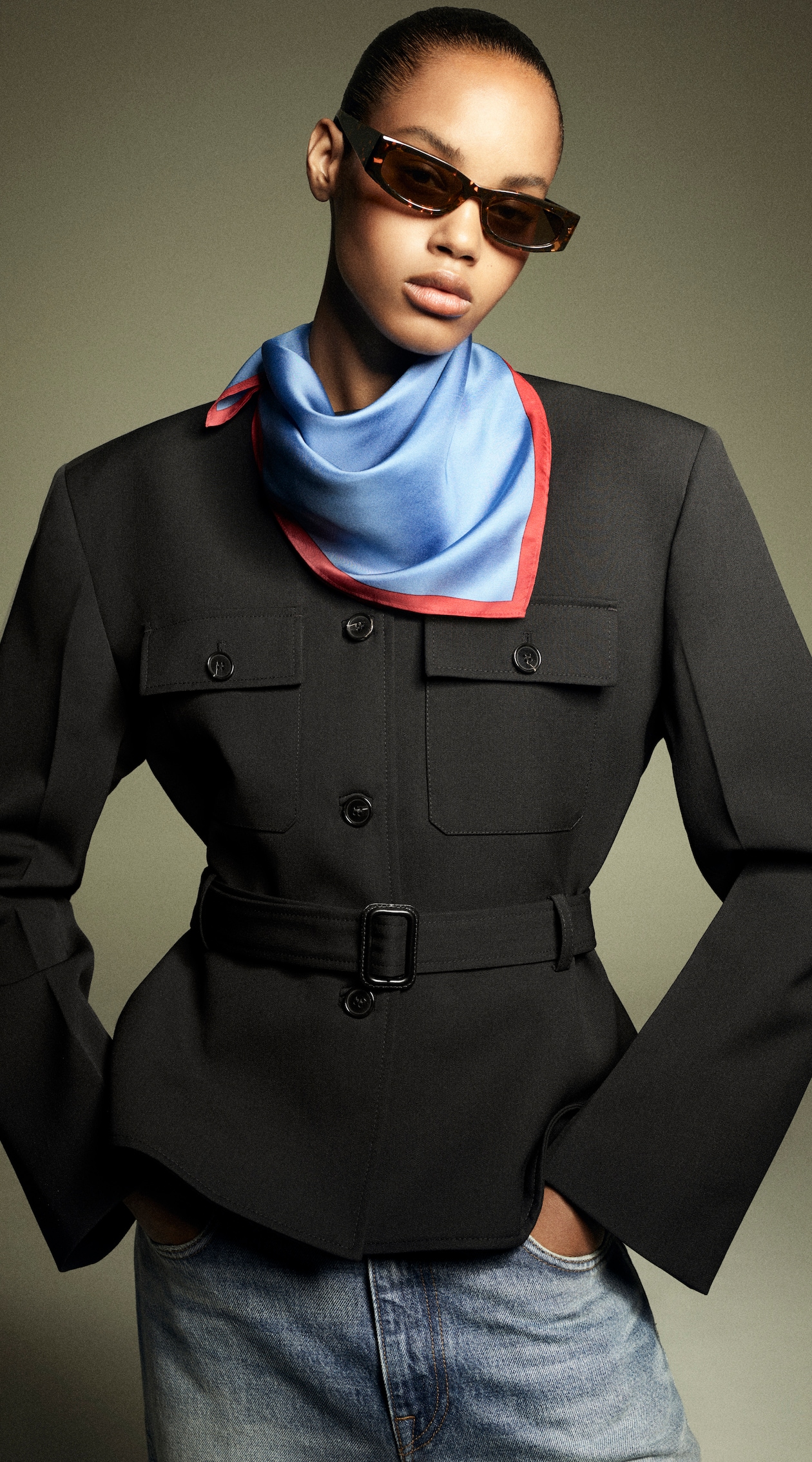 Person wearing a black belted jacket, blue and red scarf, and sunglasses, with hands in denim pockets against a neutral background.