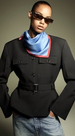 Placeholder for: Person wearing a black belted jacket, blue and red scarf, and sunglasses, with hands in denim pockets against a neutral background.