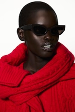 Placeholder for: A person with short hair wears black sunglasses and a vibrant red knitted turtleneck sweater, posing against a white background.