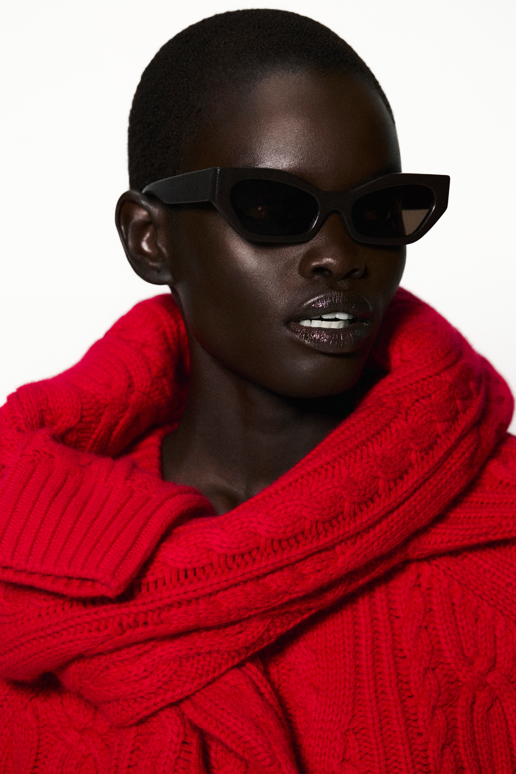 Person wearing black cat-eye sunglasses and a bright red, chunky knit sweater, posing against a plain white background.